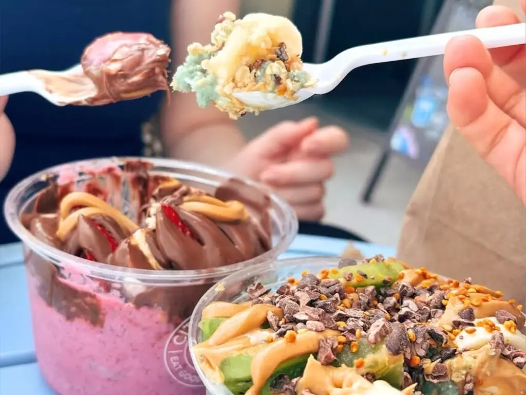 Two spoonfuls held above an açaí bowl swirled with Nutella and a green bowl topped with peanut butter, cacao nibs and bee pollen